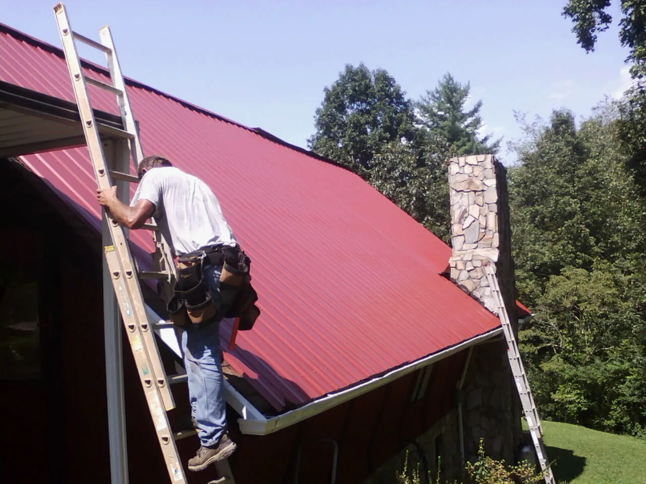 Roofer on a ladder beside a red metal roof and stone chimney in Ogdensburg